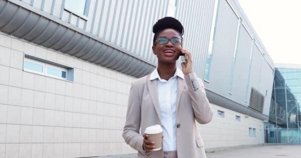 Afro-americano giovane donna d'affari felice in occhiali parlando al cellulare e in piedi all'aperto e sorseggiando bevanda calda. Bella donna in stile business che parla sul cellulare e beve caffè. — Video Stock
