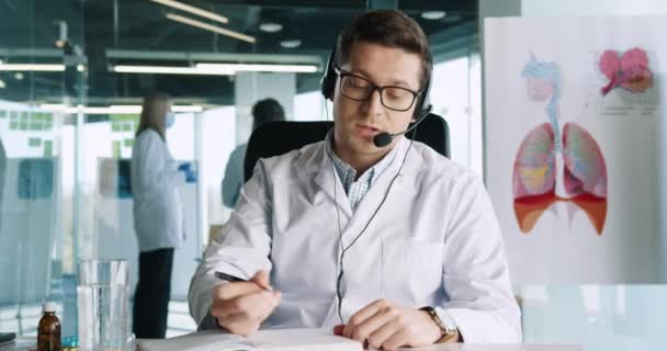 Retrato del médico varón en auriculares hablando y mirando la cámara en videollamada en línea con el paciente sentado en el gabinete en el hospital y escribiendo en el planificador que prescribe el tratamiento. Concepto de telemedicina — Vídeos de Stock