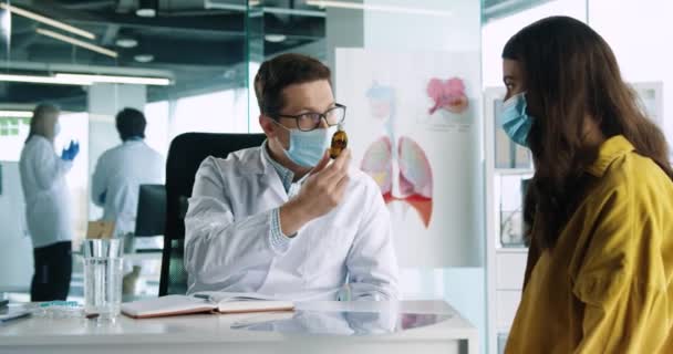 Primer plano de varón caucásico guapo joven médico en máscara médica hablando con paciente bastante femenina en consulta, explicando y mostrando la medicina. Concepto de salud. Coronavirus covid-19 — Vídeo de stock