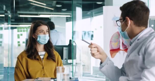 Vista lateral del guapo médico caucásico masculino con máscara médica que habla con una paciente joven en consulta en el gabinete del hospital durante la pandemia de coronavirus. Concepto de salud. De cerca. — Vídeos de Stock