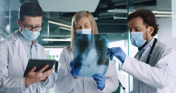 Närbild porträtt av kaukasiska manliga och kvinnliga läkare i medicinska masker talar och diskuterar röntgen scan och skriva på tablett enhet på sjukhus kontor. Kollegor arbetar på kliniken under coronavirus — Stockvideo