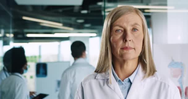 Retrato de cerca de una anciana doctora caucásica vestida con un abrigo blanco de pie en la sala de la clínica y mirando a la cámara. Médicos de razas mixtas con máscaras trabajando en el fondo. Concepto de Coronavirus — Vídeo de stock