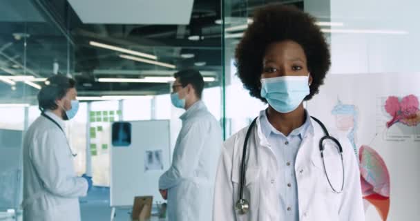 보호 마스크를 쓰고 클리닉에 서서 카메라를 보고 있는 젊은 아프리카 여성 의사의 사진을 클로즈업 합니다. 백인 의사들은 배경 연구를 하고 있어요. 코로나 바이러스의 개념 — 비디오