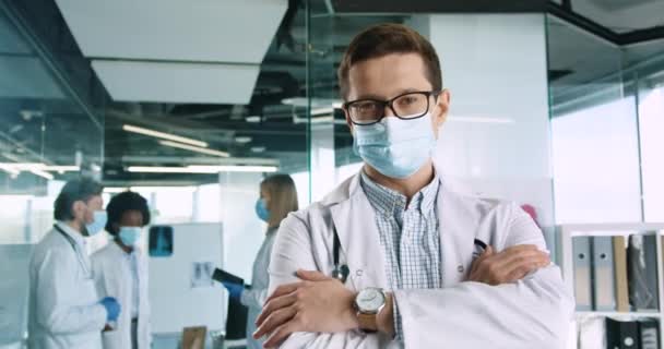 Close up of Caucasian joyuful handsome young male professional medical practitioner in glasses wearing mask standing in clinic and looking at camera. Mixed-races doctors on background. Covid19 concept — Stock Video