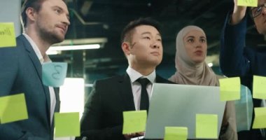 Modern ofiste çalışan ve cam tahtaya yazı yazan birçok melez erkek ve kadın profesyonel iş arkadaşı şirket kurma projesini tartışıyorlar. Takım çalışması konsepti. Girişimcilik