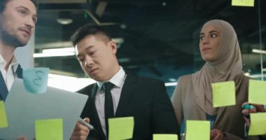 Modern ofiste çalışan, cam tahtaya yazı yazan, dizüstü bilgisayara bakan ve iş kurma projesini tartışan birçok melez erkek ve kadın pazarlamacıyı kapatın. Takım çalışması konsepti