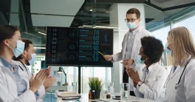 Tıp maskeli genç erkek doktor, sağlık sektöründe çalışan iş arkadaşlarıyla Coronavirus hastalıkları istatistikleri ve hastanede olumlu sonuçlar hakkında konuşurken meslektaşları el çırpıyor. Covid 19