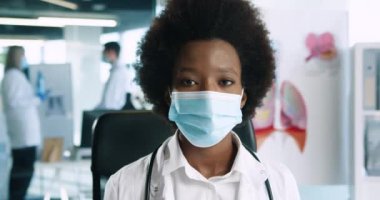 Tıbbi maske takmış Afro-Amerikalı mutlu kadın doktorun yakın plan portresi iş yerinde kameraya bakıyor. İş yerindeki sağlık uzmanı. Coronavirus enfeksiyonu konsepti