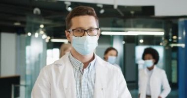 Sağlık maskeli beyaz erkek doktorun yakın plan portresi. Çalışma yerinde kameraya bakıyor. Klinikte duruyor ve arka planda iş arkadaşları var. Coronavirus enfeksiyonu konsepti