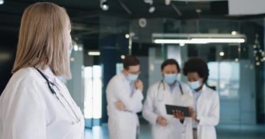 Kafkas kıdemli tıbbi maskeli kadın doktorun yakın plan portresi hastanede kameraya bakıyor. Sağlık uzmanları arka planda tablet kullanıyor. Coronavirus konsepti