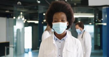 Afro-Amerikalı mutlu kadın sağlık uzmanının yakın plan portresi tıbbi maske uzmanı arka planda konuşan doktorlarla çalışırken kameraya bakıyor. Coronavirus enfeksiyonu. Covid-19 konsepti