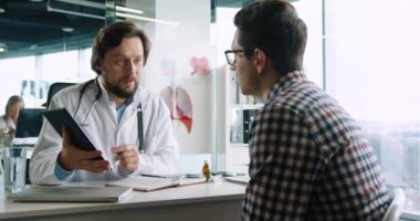 Steteskoplu yakışıklı erkek doktorun portresini yakından çek. Hastanedeki masada otururken hastayla görüşürken tablete bak ve daktilo yaz. Sağlık hizmetleri kavramı