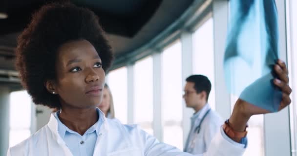 Close up portrait of African American young beautiful female professional healthcare specialist standing in modern hospital and looking at X-ray scan. Doctors talking on background — Stock Video