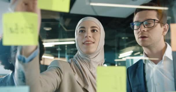 Close up of mixed-races male and female professional working in modern office. Beautiful Arabian female writing on glass board in cabinet and speaking with colleagues. Business concept — Stock Video