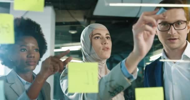 Close up of multi-ethnic male and female professional managers working in modern office. Beautiful Arabian female writing on glass board and talking to team while man typing on tablet Business concept — Stock Video