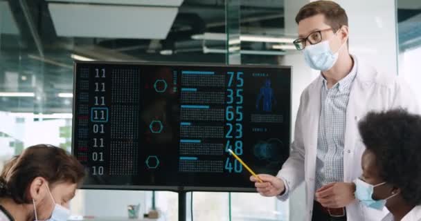 Крупним планом чоловічий лікар у масці обговорює з багатонаціональними медичними працівниками статистику коронавірусної хвороби в лікарні, дивлячись на цифровий монітор. Жінка друкує на планшеті. Вірус коїд-19 — стокове відео