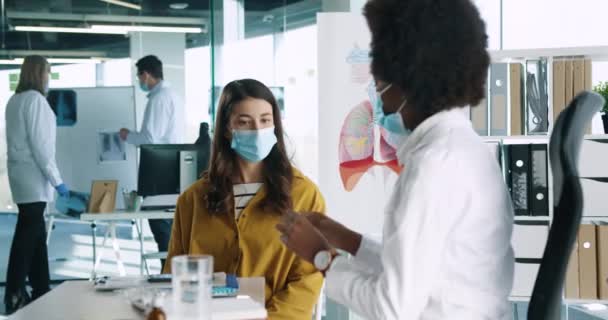 Retrato de cerca de la médica afroamericana en máscara médica sentada en el trabajo explicando a la mujer el tratamiento de la medicina del paciente de cronavirus en la consulta médica Concepto de infección Covid-19 — Vídeo de stock