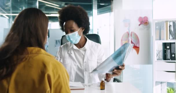 Nahaufnahme Porträt einer jungen afroamerikanischen Ärztin in medizinischer Maske, die am Arbeitsplatz sitzt und dem Röntgenbild zusieht, das der Patientin das Ergebnis und die Diagnose erklärt. Coronavirus covid-19-Konzept — Stockvideo