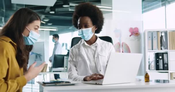 Close up retrato de jovem afro-americana bonita médico em máscara médica sentar no local de trabalho falando em consulta com paciente do sexo feminino. Coronavirus covid-19. Conceito de centro médico — Vídeo de Stock