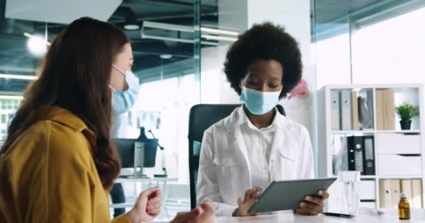 Retrato de una bonita médica afroamericana con máscara médica sentada en el trabajo en la clínica, hablando con una paciente durante la consulta médica y escribiendo en un dispositivo de tableta. Concepto de Coronavirus — Vídeos de Stock