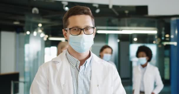 在工作场所，戴着医疗面罩的白人快乐男医生的近照，与同事一起站在诊所里，看着镜头，心情很好。Coronavirus感染概念 — 图库视频影像