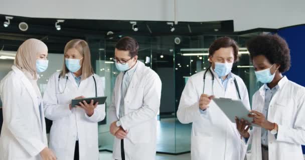 Wielu etnicznych lekarzy płci męskiej i żeńskiej pracownicy służby zdrowia w maskach medycznych dyskutują o zakażeniu koronawirusem, pisząc na tabliczce stojącej w klinice. Koncepcja portretu. Pandemia wywołana przez wirus Covid-19 — Wideo stockowe