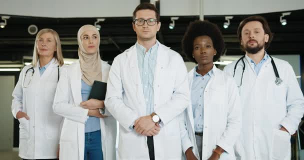 Ritratto di giovani e anziani medici caucasici e afroamericani in camici medici in piedi in laboratorio in ospedale a guardare la telecamera. Squadra degli operatori sanitari in clinica. Concetto di medicina — Video Stock