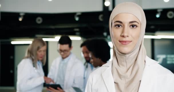 Close up portret młodej szczęśliwej pięknej kobiety lekarz stojący w szpitalu w płaszczu medycznym patrząc na aparat w dobrym nastroju z pracownikami służby zdrowia za pomocą tabletu i rozmowy na tle — Wideo stockowe