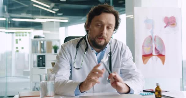 Porträt eines schönen jungen erwachsenen männlichen Arztes mit Stethoskop, der in der Klinik am Arbeitsplatz sitzt, spricht und medizinische Beratung gibt. Coronavirus-Pandemie. Krankenhauskonzept — Stockvideo