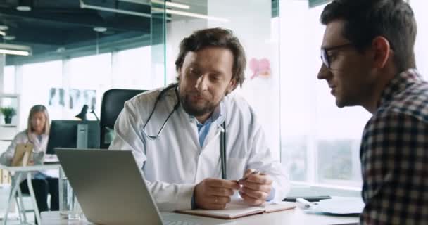 Primer plano retrato de guapo médico masculino con estetoscopio sentado en el escritorio en el hospital en el trabajo mirando a la computadora portátil y hablando con el paciente en consulta. Concepto de salud. Asesoramiento médico — Vídeo de stock