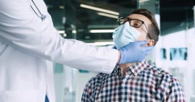 Gözlüklü, tıbbi maske takan, doktor randevusu için hastane ofisinde oturan erkek hastayı kapatın. Doktor klinikteki genç hastayı muayene ediyor. Sağlık hizmetleri kavramı