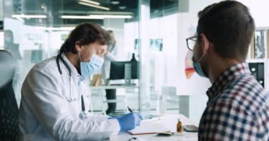 Tıbbi maske takan erkek doktorların hastane masasında oturan genç bir hastayla görüşmelerini kapatın. Doktor randevusundaki genç adamın arkası. Coronavirus konsepti