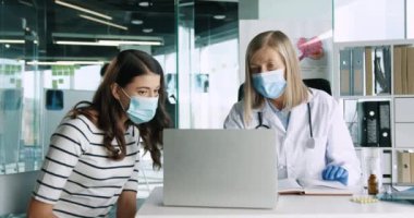 Dizüstü bilgisayara ve daktiloya bakan kıdemli kadın doktorla muayenede klinikte oturan genç bir hastanın portresini yakından çek. Coronavirus konsepti. Kapsamlı salgın