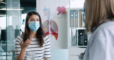 Tıbbi maske takan, kıdemli kadın doktorla istişare için doktor ofisinde oturan genç ve güzel bir hastaya yakından bakın. Koronavirüs salgını. Portreyi kapat. Covid-19 tedavisi