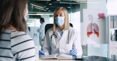 Klinikteki kabinede oturan ve genç beyaz kadın hastayla görüşen tıbbi maskeli yaşlı kadın doktor portresi. Erkek doktorlar arka planda konuşuyor. Klinik kavramı