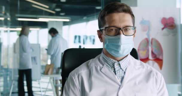 Close up retrato de belo jovem médico em óculos sentado no escritório do hospital no trabalho, olhando para a câmera e sorrindo. Trabalhador de saúde no gabinete. Conceito de profissão médica — Vídeo de Stock