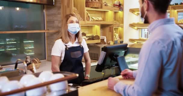 Trabajadora de panadería caucásica en máscara médica se para en el trabajo y habla con un cliente varón que vende pan. Concepto de pequeña empresa. Mujer en delantal en el mostrador de la panadería. Concepto alimenticio — Vídeo de stock