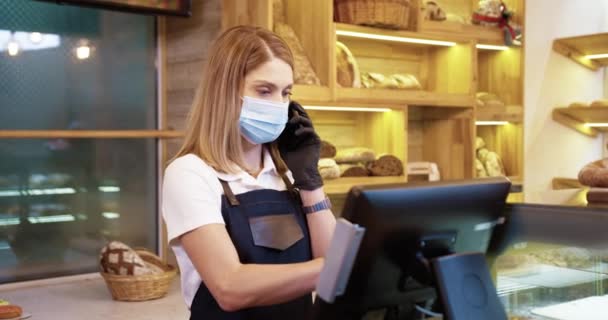 Retrato de la vendedora caucásica en máscara hablando por teléfono móvil en la cafetería y tocando en la computadora tomando la orden del cliente a través del teléfono celular. Panadería, dueño de una pequeña empresa, concepto de industria alimentaria — Vídeo de stock