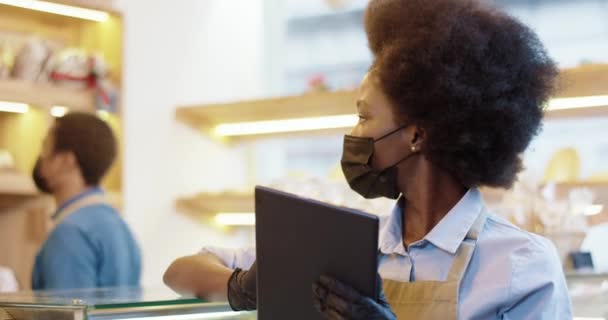 Primer plano de la mujer bonita afroamericana vendedora con máscara negra y guantes de pie en la cafetería escribiendo en la tableta en el lugar de trabajo en la panadería, mientras que su compañero de trabajo masculino trabajando en el fondo de la panadería — Vídeo de stock