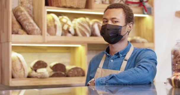 Primer plano retrato de afroamericano joven vendedor guapo en la máscara de pie en panadería en el mostrador en la panadería, mirando a la cámara de buen humor. Dueño de una pequeña empresa. Concepto Baker — Vídeos de Stock