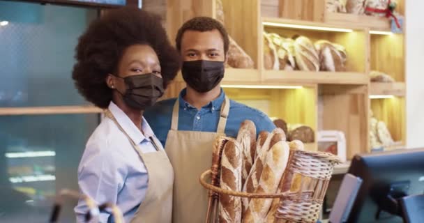 Retrato da alegre família jovem afro-americana casal padeiros em máscaras de rosto segurando cesta com pão fresco em pé em pequena própria padaria em quarentena. Negócios familiares. Conceito de pão — Vídeo de Stock