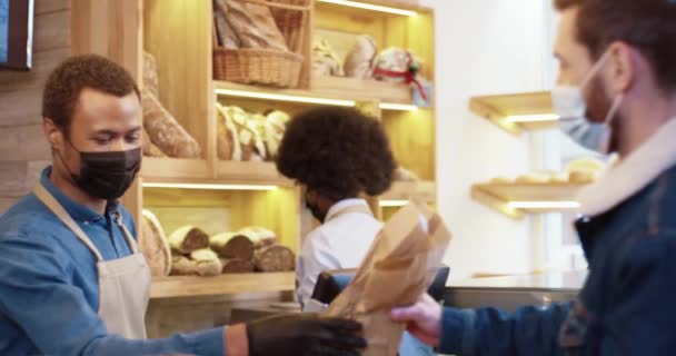 Close up de vendedor masculino afro-americano em máscara e luvas vendendo pão fresco na padaria enquanto colega trabalhando atrás dele. Cliente que paga com cartão de crédito no dispositivo. Conceito de Bakehouse — Vídeo de Stock