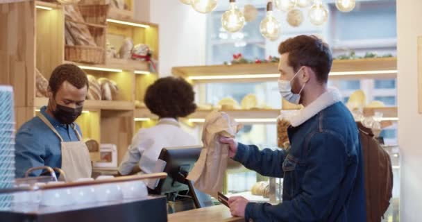 Vista lateral del joven vendedor afroamericano en máscara y guantes que venden pan fresco en la panadería mientras una colega trabaja detrás de él. Cliente masculino pagando con tarjeta de crédito en panadería — Vídeo de stock