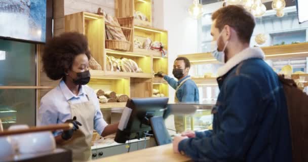 Boční pohled na mladou afro-americkou prodavačku v masce prodávající pečení v pekárně při objednávce od kavkazského klienta stojícího za pultem pekařství na počítači — Stock video