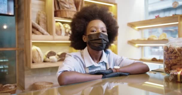 在面包店柜台旁的面包店里工作的戴着面具的非洲裔美国漂亮女卖家的近照，看着相机，面带微笑。小企业主概念 — 图库视频影像