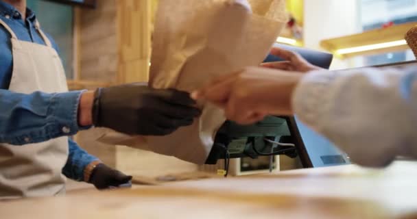 Närbild av afroamerikanska manliga säljare i ansiktsmask och skyddande svarta handskar som säljer bakat färskt bröd i bageriet butik. Klienten betalar med kreditkort på enheten köper bakning. Affärsidé — Stockvideo