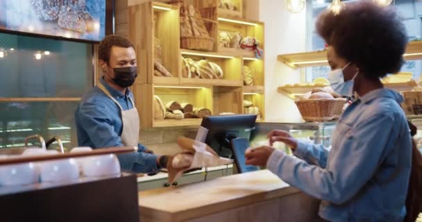 Vista lateral del vendedor masculino afroamericano en mascarilla que vende pan fresco horneado en la panadería al cliente bastante femenino. Mujer pagando con tarjeta de crédito comprando panadería. Concepto de compras — Vídeos de Stock