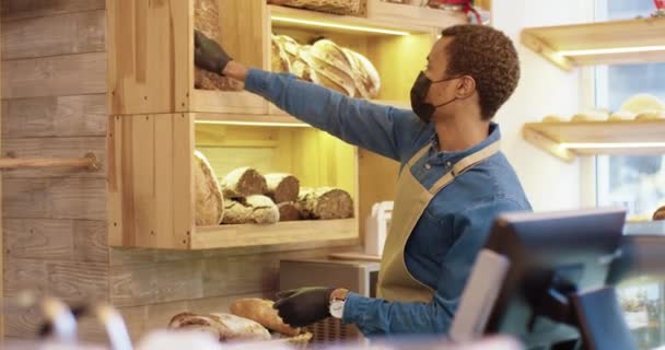 Sisi tampilan. Pekerja muda Afrika Amerika mengenakan masker wajah dan celemek berdiri di toko roti dan menempatkan roti panggang segar di rak-rak. Pria pemilik toko roti bekerja sendirian di warung kopi kecil — Stok Video