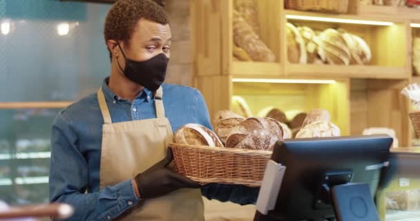 Primer plano de guapo joven afroamericano joven panadero trabajador en la máscara de la cara de pie en la panadería y la celebración de la cesta con pan recién horneado. Propietario de panadería mirando a la cámara y sonriendo — Vídeo de stock