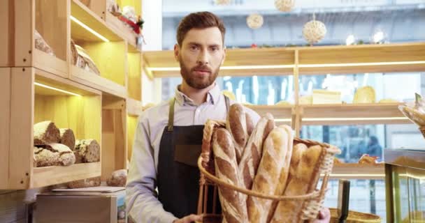 パン屋さんに立つエプロン姿や焼きたてのパンを手にしたバスケットを持ち、カメラを見て微笑むハンサムな若い白人男性販売員の姿。中小企業コンセプト — ストック動画